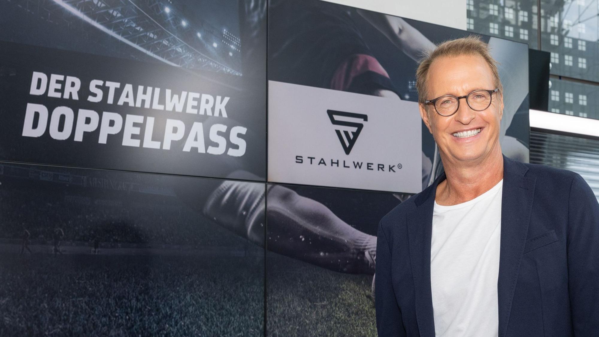 Sportmoderator Florian König lächelt vor dem „Doppelpass“-Logo in die Kamera.