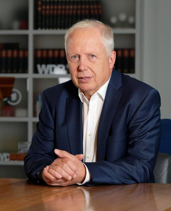 WDR-Intendant Tom Buhrow setzt in einem blauen Anzug an einem Tisch und schaut in die Kamera.