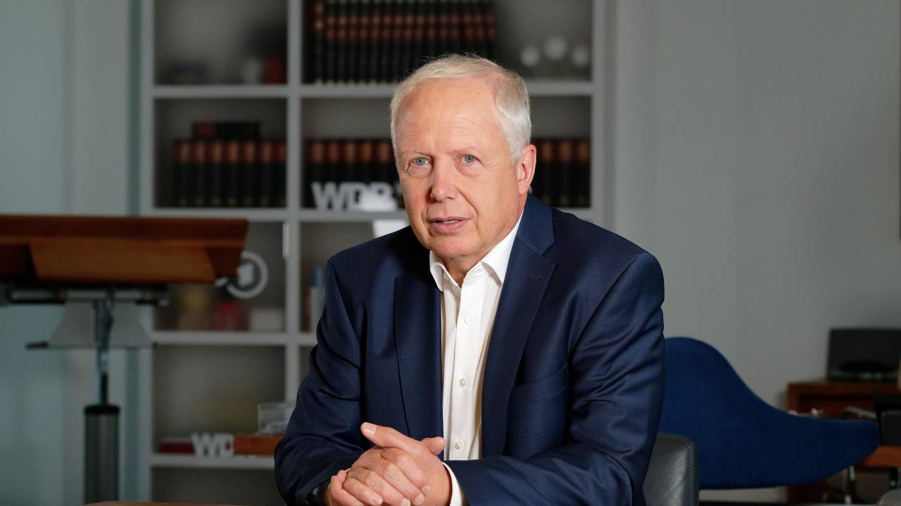 WDR-Intendant Tom Buhrow setzt in einem blauen Anzug an einem Tisch und schaut in die Kamera.