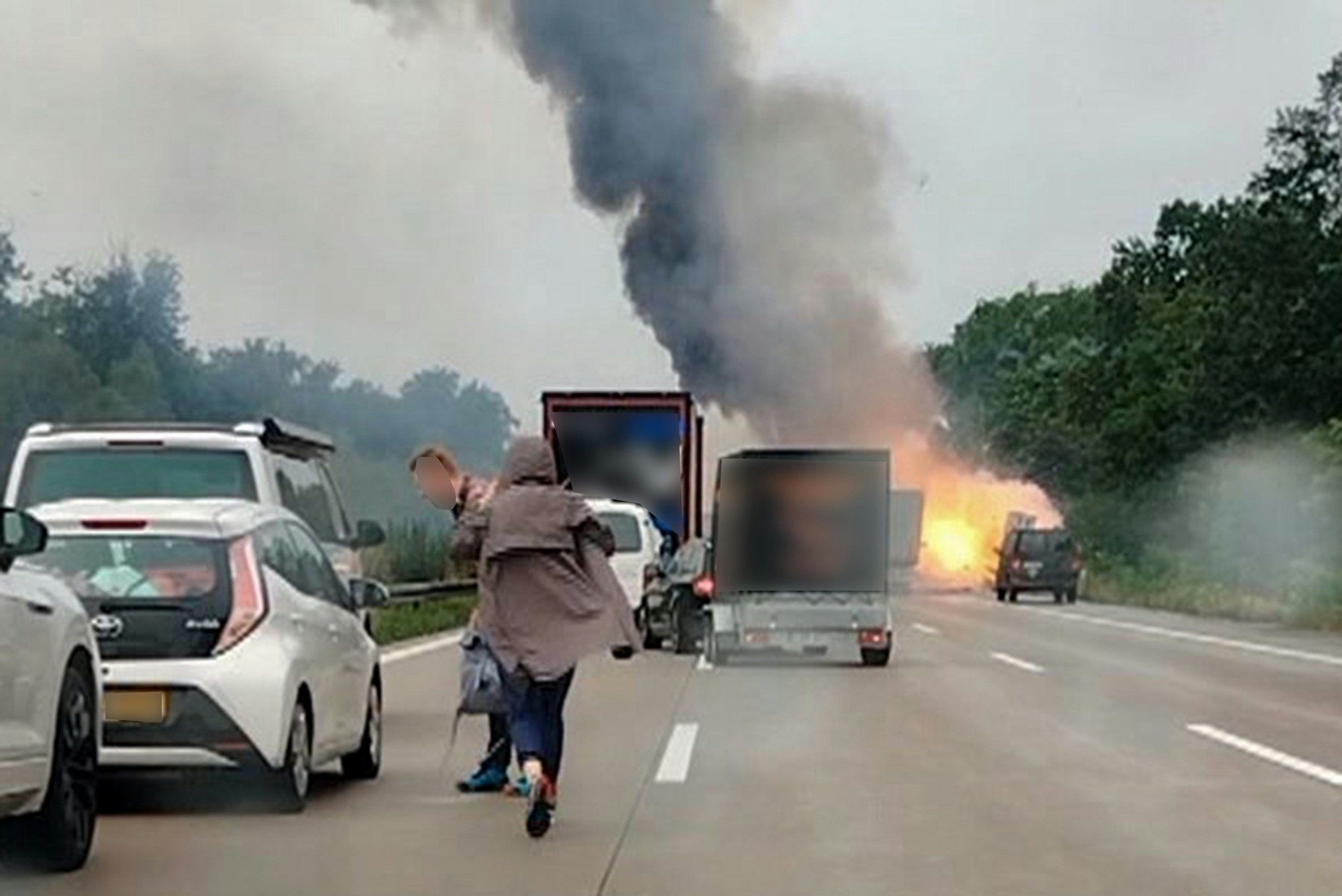 Autofahrer und -fahrerinnen verlassen ihre Fahrzeuge, während im Hintergrund ein LKW in Flammen.
