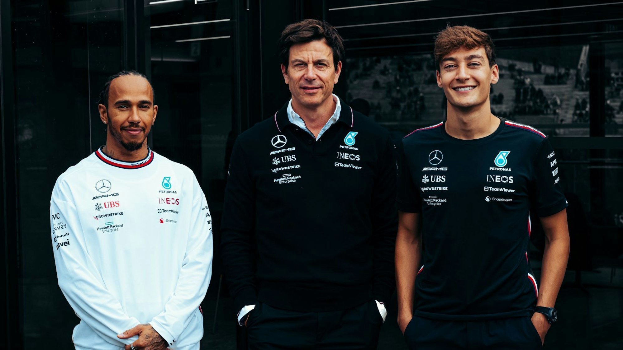 Lewis Hamilton (li.), Toto Wolff (Mitte) und George Russell (re.) posieren für ein Foto und lächeln.