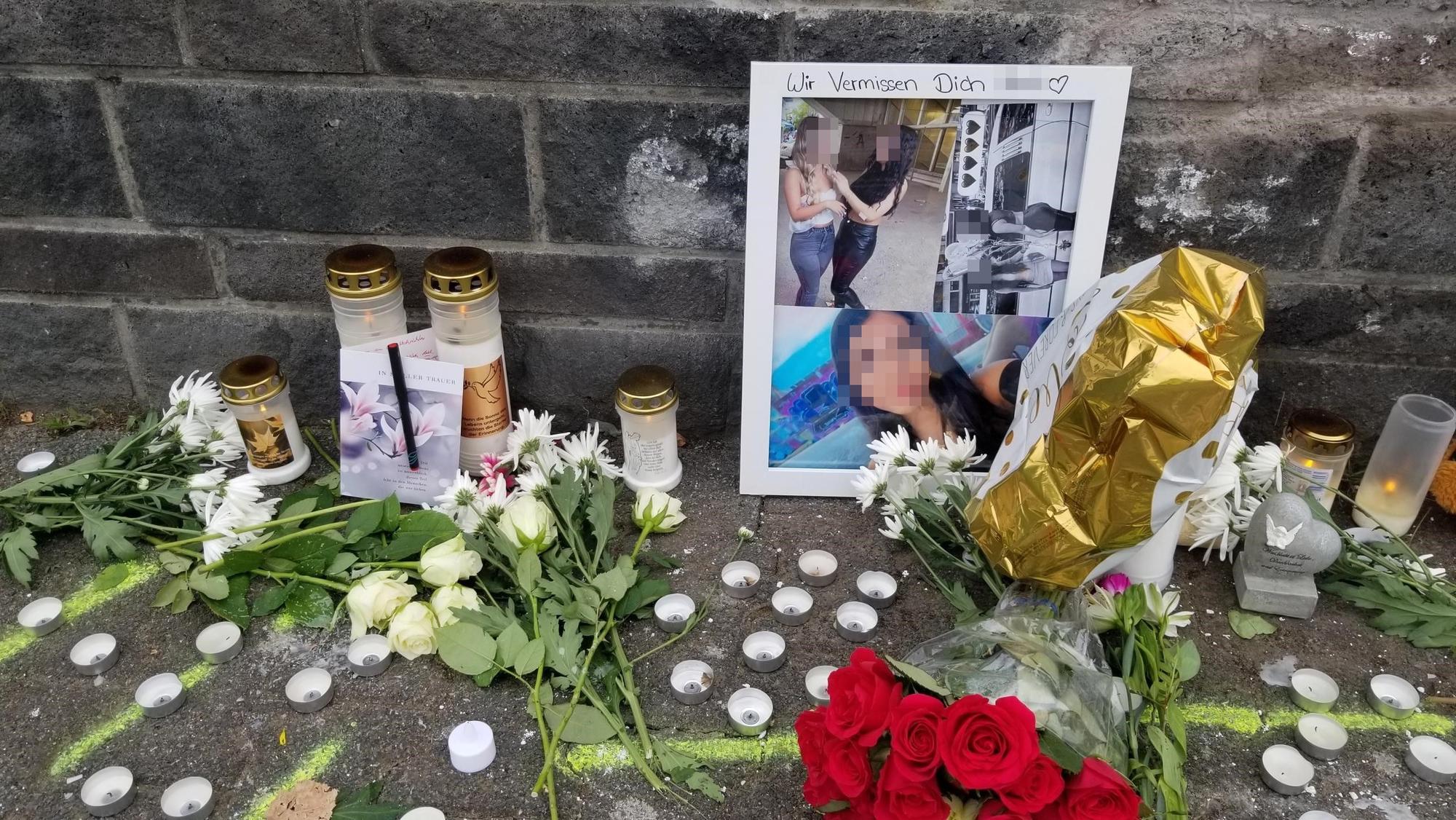Vor einer Mauer stehen Fotos und Kerzen, liegen Blumen.