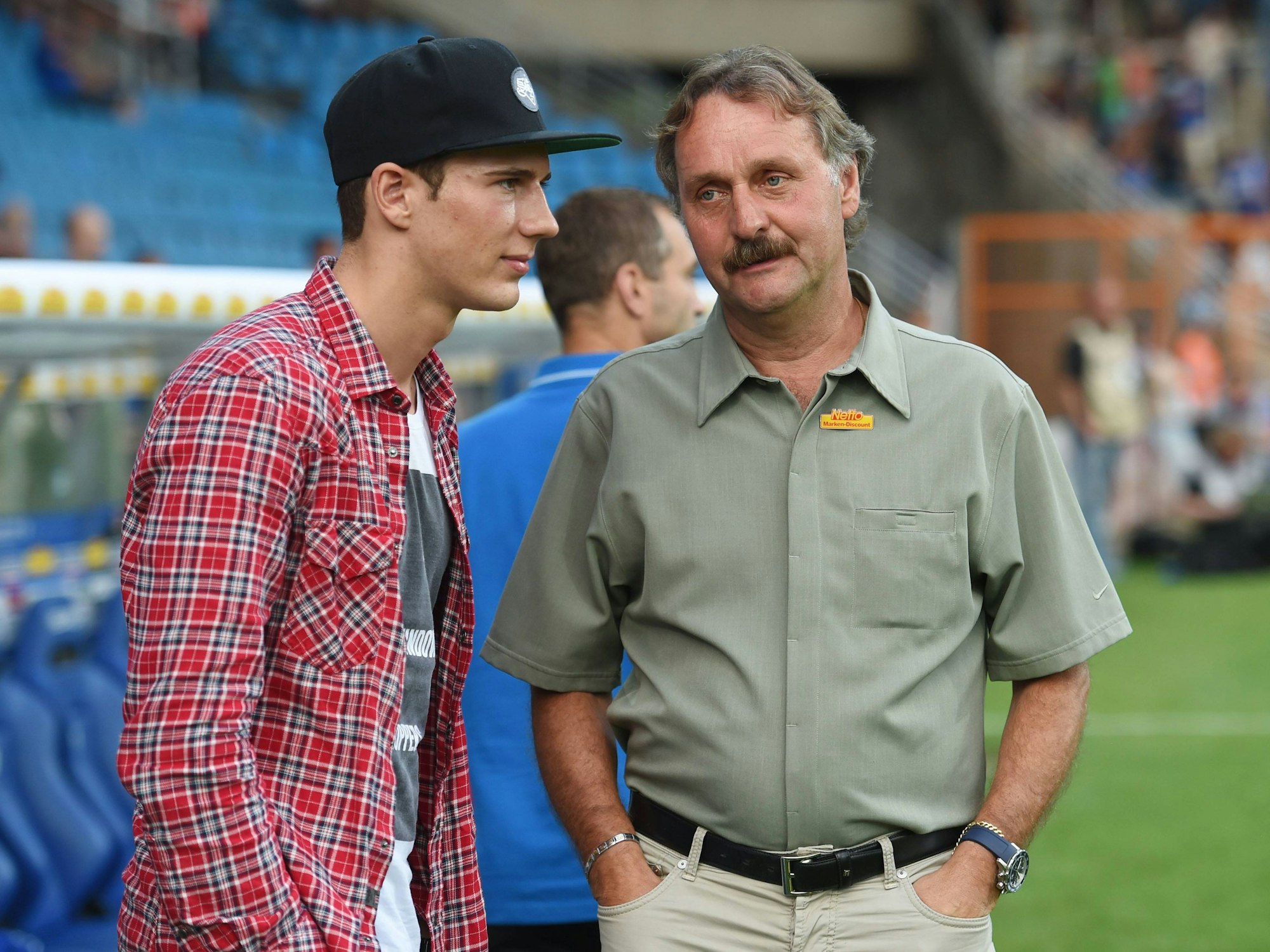 Peter Neururer hat die Hände in den Hosentaschen. Neben ihm steht Leon Goretzka. Er trägt eine Kappe.