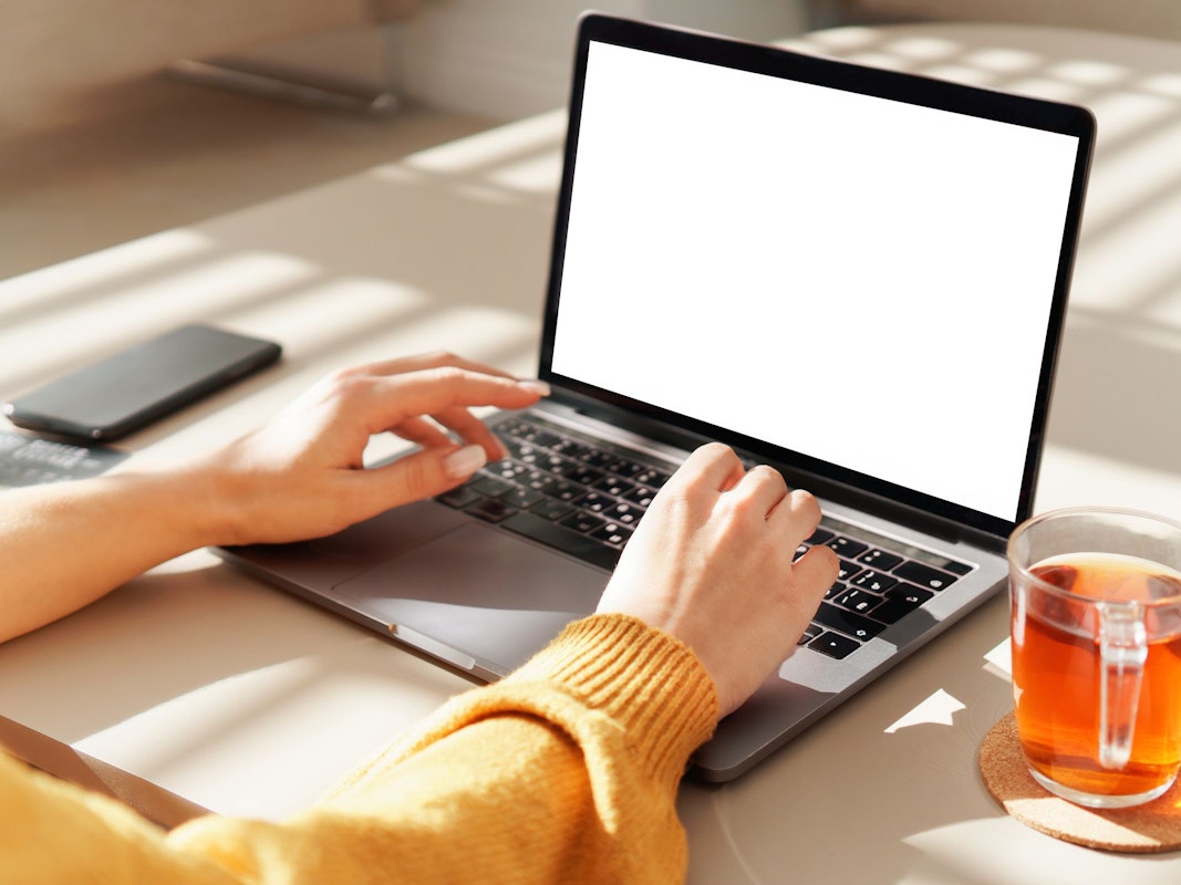 Eine Frau tippt auf einem Notebook. Neben ihr steht ein Glas mit Tee.