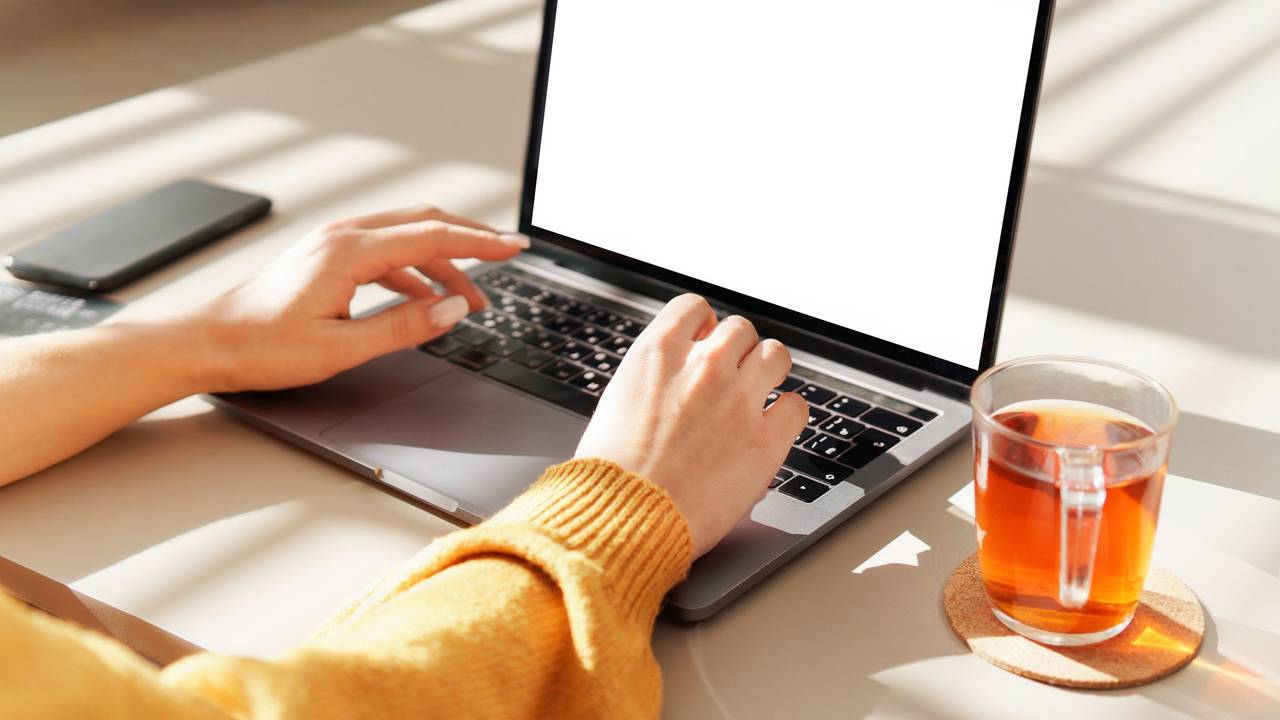 Eine Frau tippt auf einem Notebook. Neben ihr steht ein Glas mit Tee.