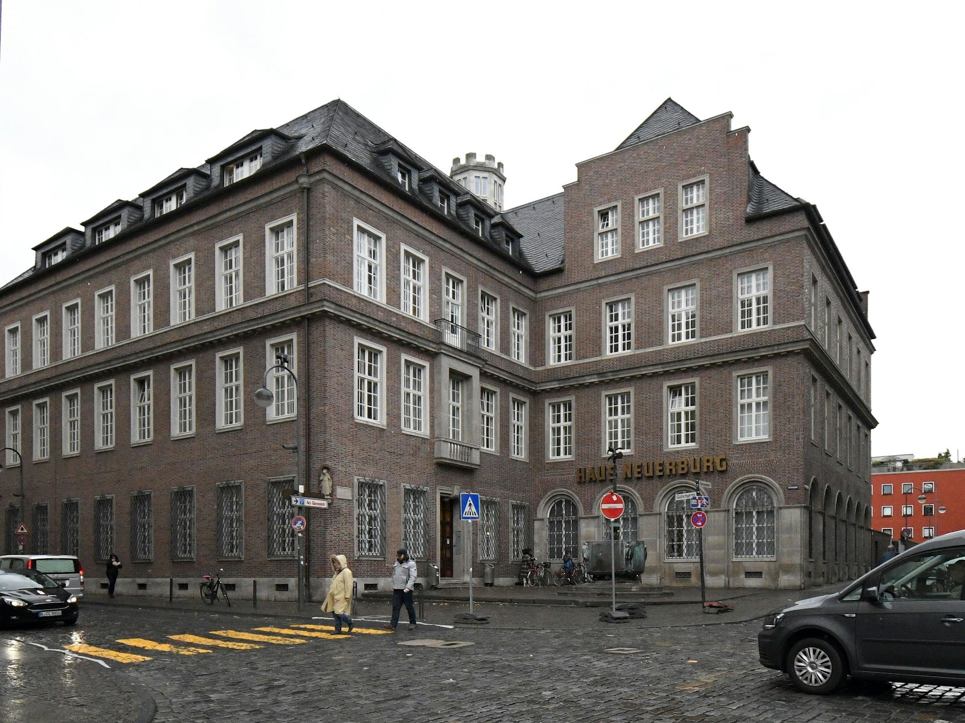 Das Haus Neuerburg in Köln bei Regen. Mehrere Menschen und Autos laufen bzw. fahren vorbei.