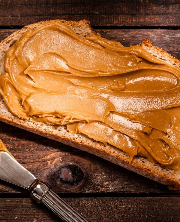 Auf dem Foto sieht man ein geschmiertes Brot mit Erdnussbutter. Daneben liegen das Glas und ein Messer.