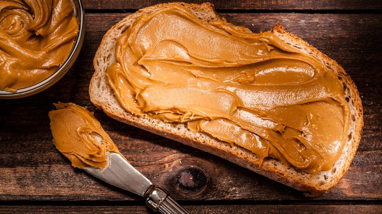 Auf dem Foto sieht man ein geschmiertes Brot mit Erdnussbutter. Daneben liegen das Glas und ein Messer.