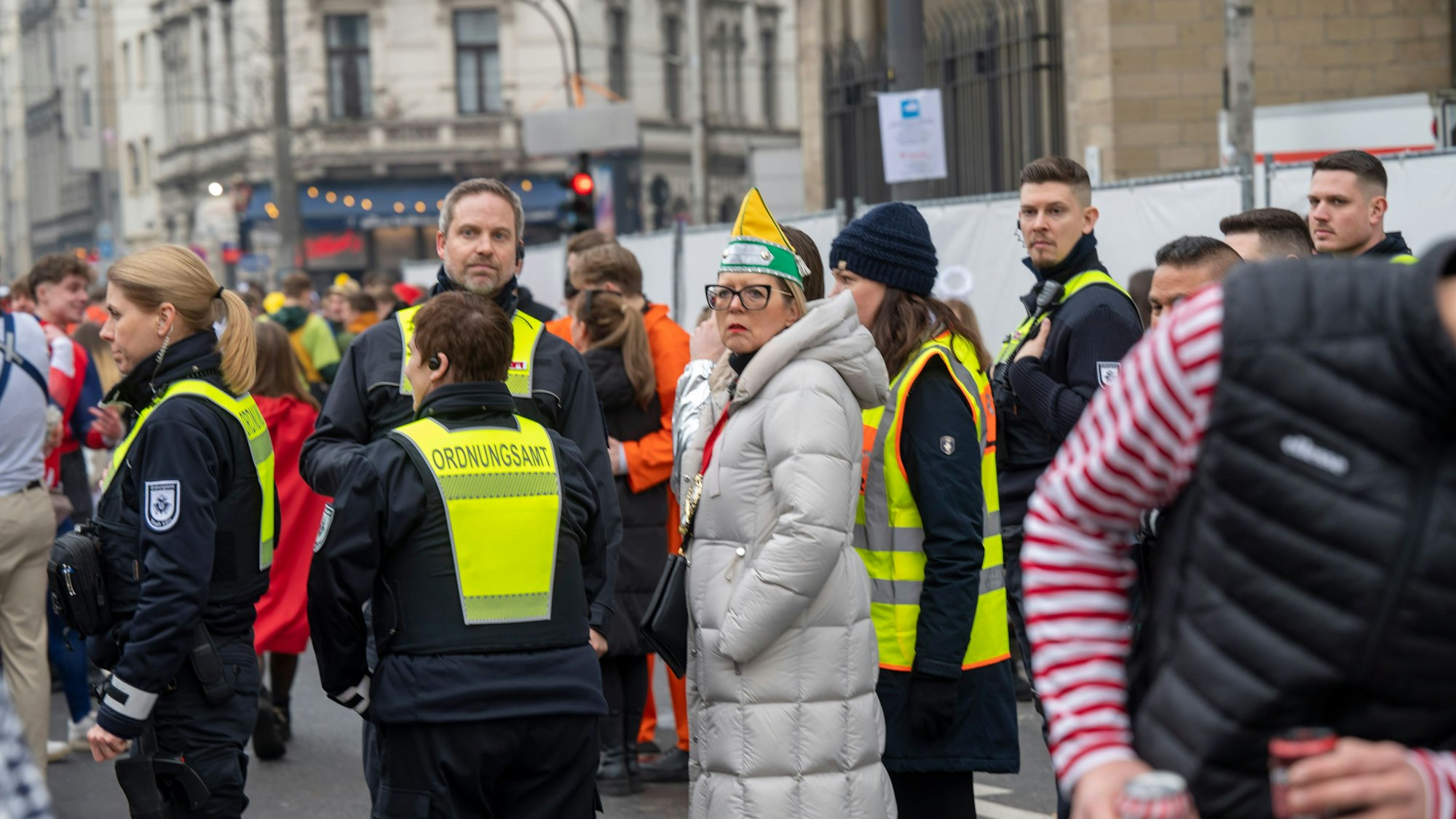 Eine Frau mit Narrenkappe steht mit Einsatzkräften des Ordnungsamts auf einer Straße in Köln.
