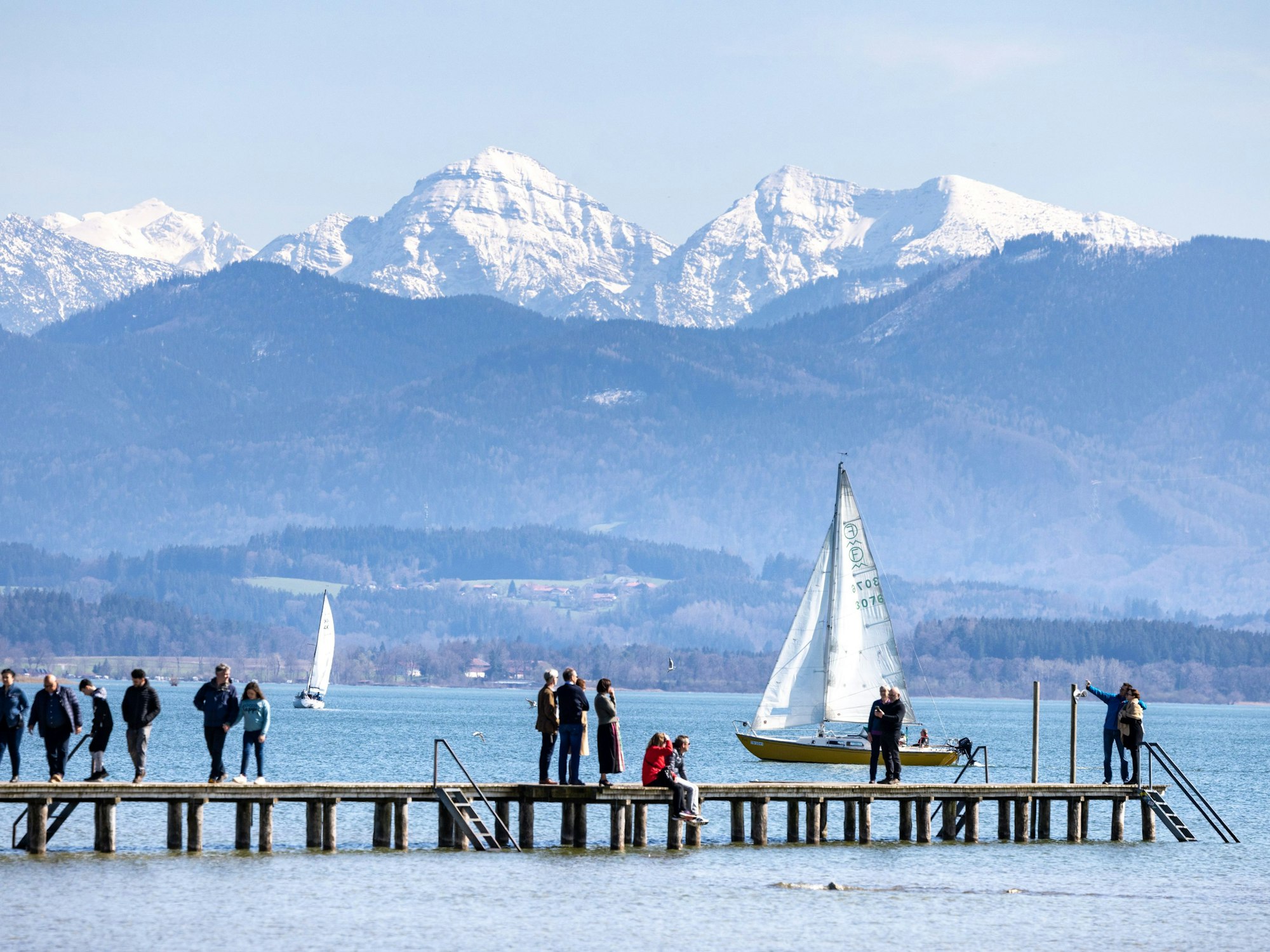 Spaziergänger und Spaziergängerinnen stehen auf einem Steg am Chiemsee, auf dem Segelboote fahren, hier im April 2023 in Bayern.