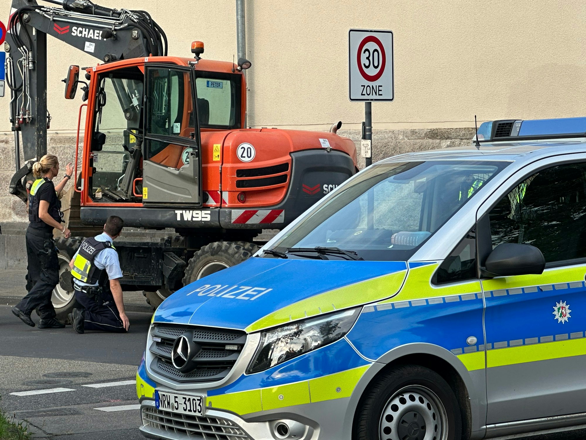 Ein Polizist hockt vor einem Bagger, eine Polizistin steht daneben. Vor dem Bagger steht quer ein Polizeiwagen.