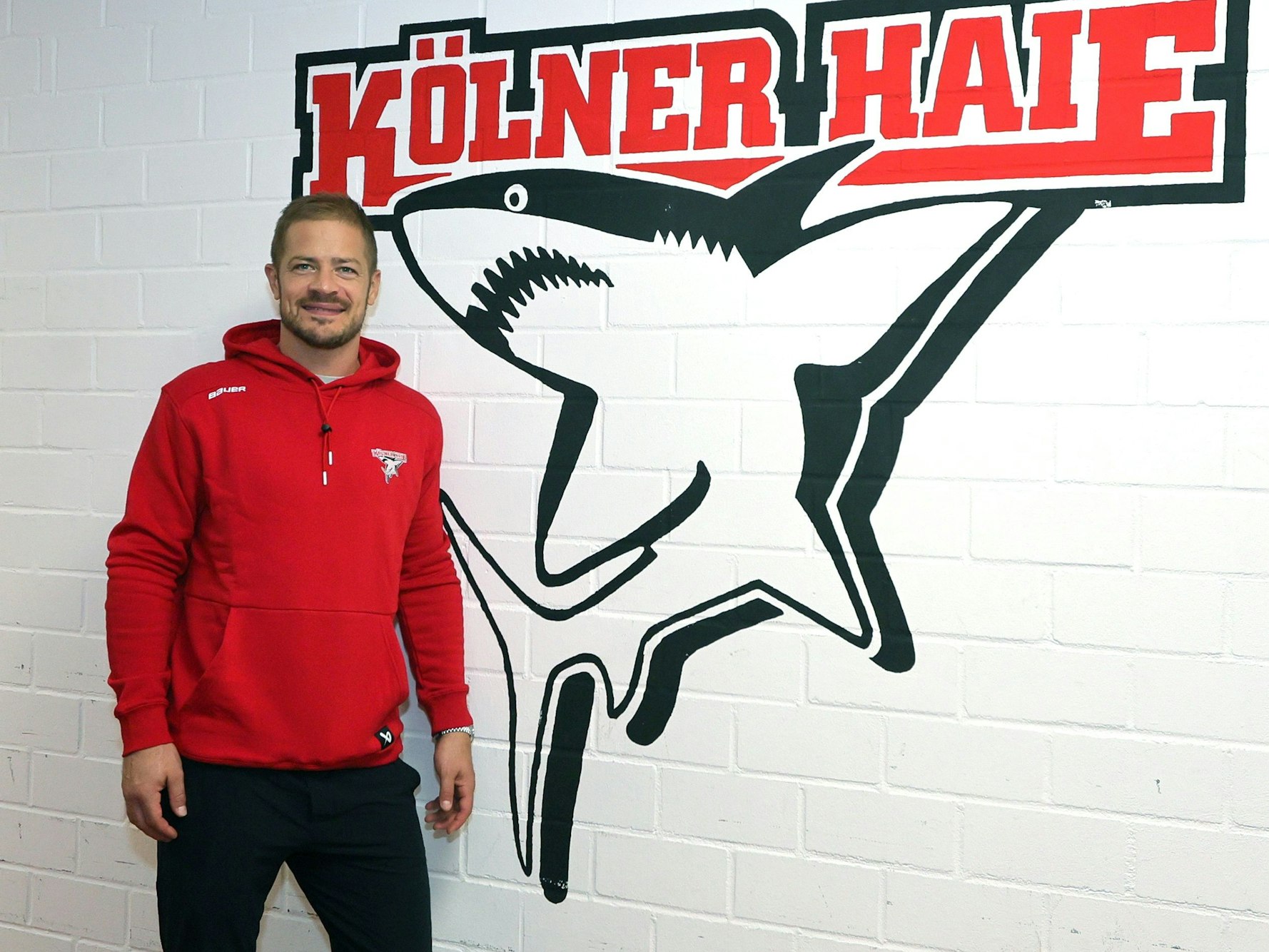 Moritz Müller (Haie) steht vor dem Logo des KEC in der Arena-Kabine.