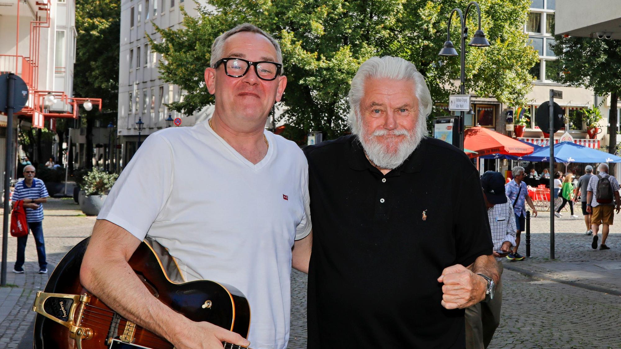 King Size Dick (r.) und JP Weber stehen in einer Kölner Fußgängerzone, halten sich im Arm und grinsen in die Kamera.