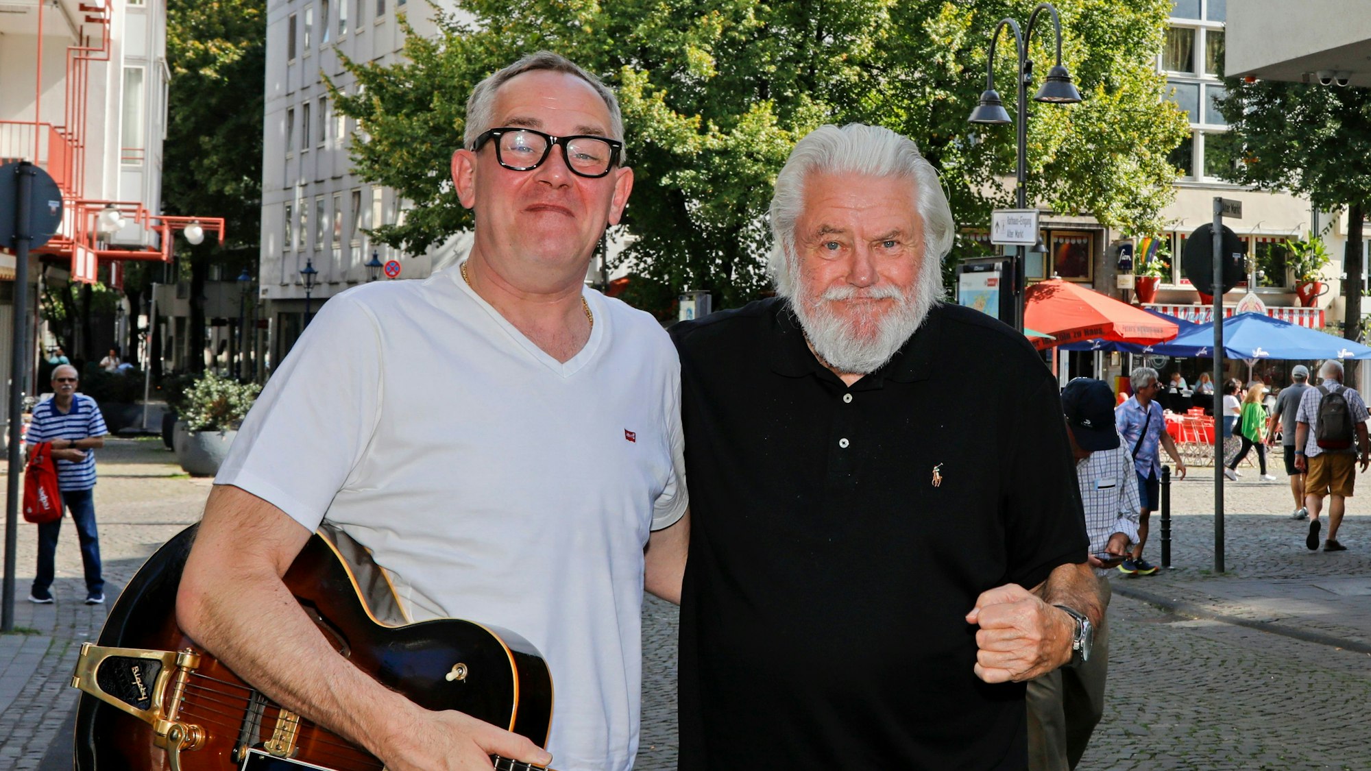 King Size Dick (r.) und JP Weber stehen in einer Kölner Fußgängerzone, halten sich im Arm und grinsen in die Kamera.