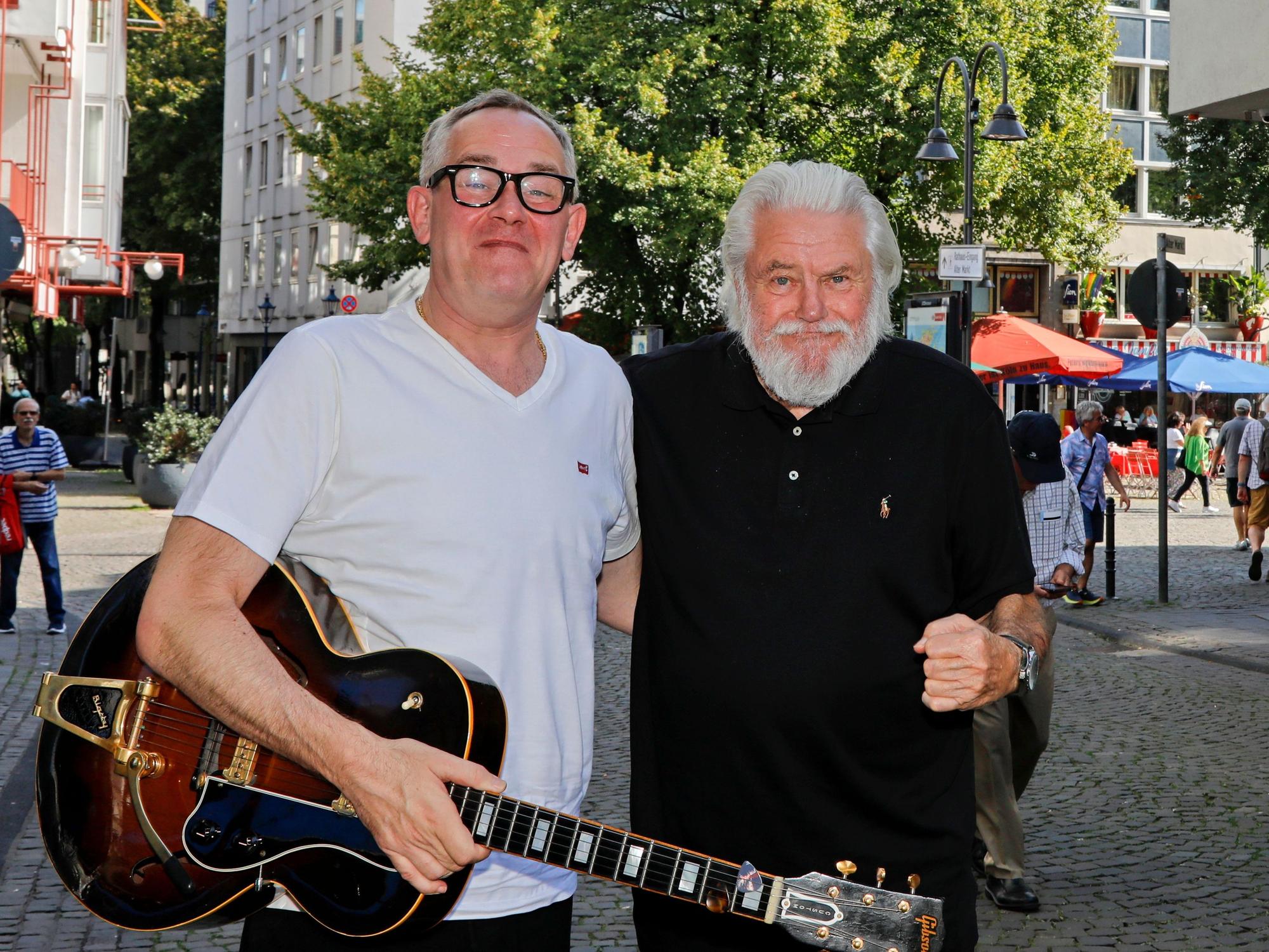 King Size Dick (r.) und JP Weber stehen in einer Kölner Fußgängerzone, halten sich im Arm und grinsen in die Kamera.