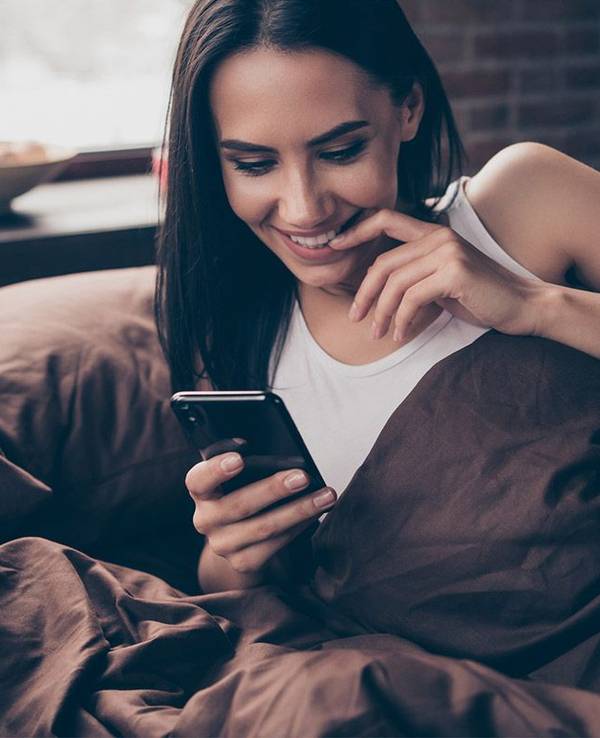 Frau im Bett schaut freudig gespannt auf ihr Handy.