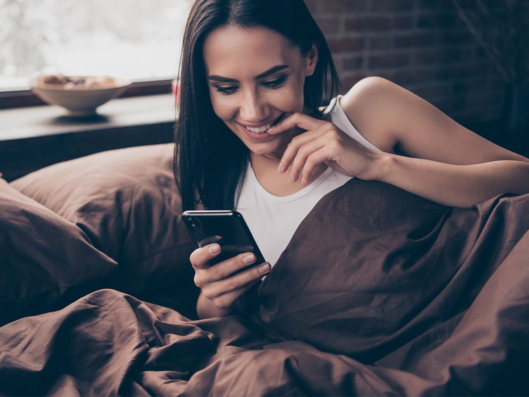 Frau im Bett schaut freudig gespannt auf ihr Handy.
