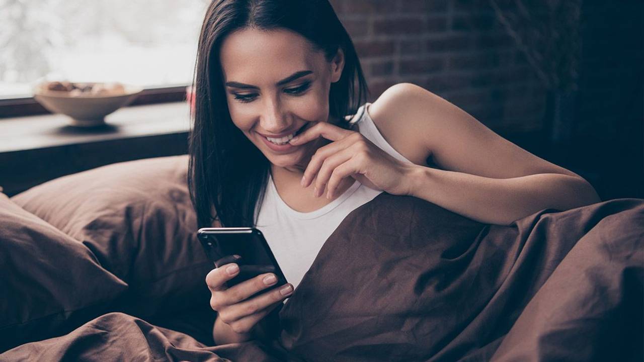 Frau im Bett schaut freudig gespannt auf ihr Handy.