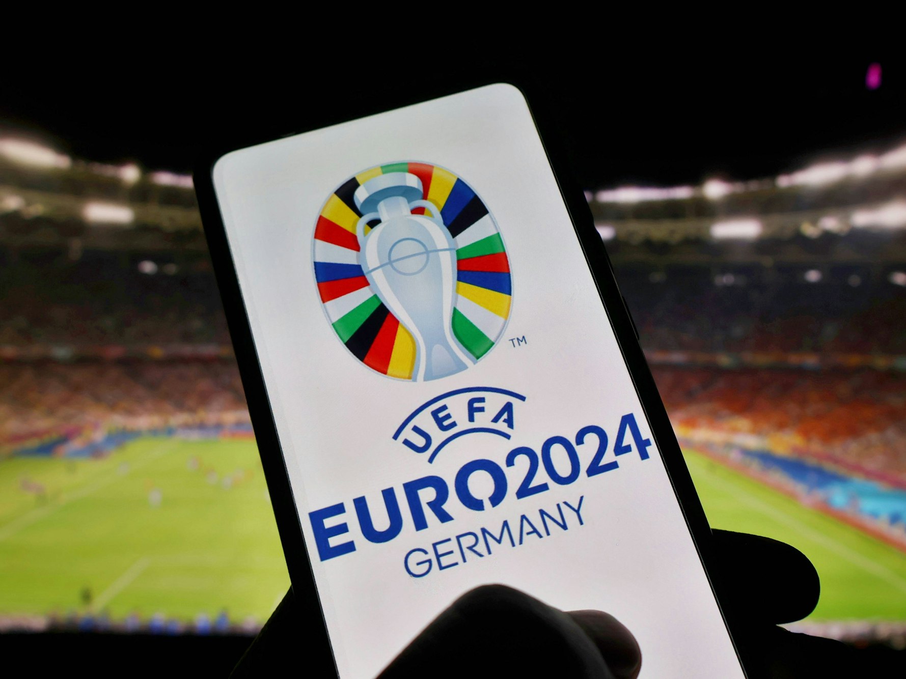 Das Display eines Handys zeigt das Logo der EM 2024 in Deutschland. Im Hintergrund ist der Innenraum eines Stadions zu
