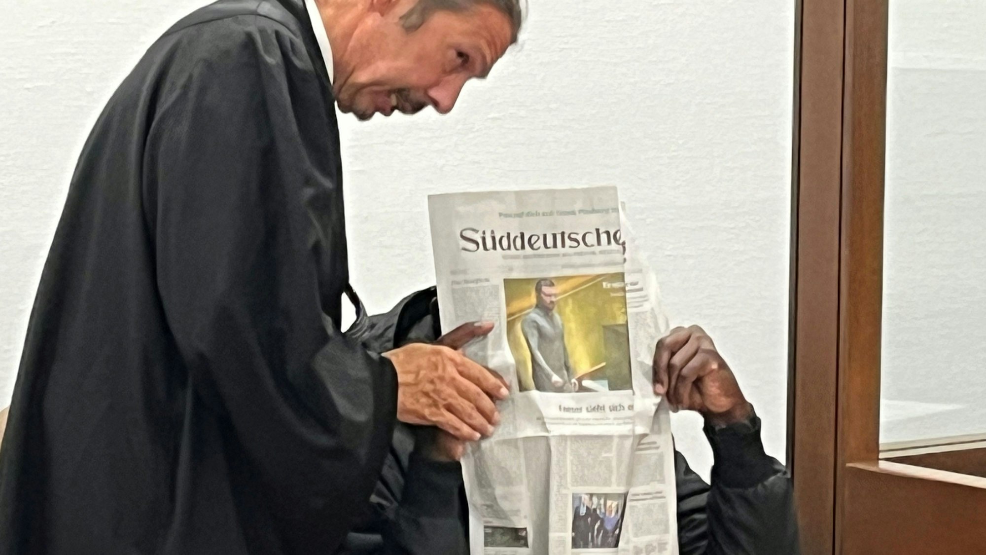 Der sitzende Beschuldigte hält sich eine Zeitung vor das Gesicht, neben ihm steht sein Anwalt.