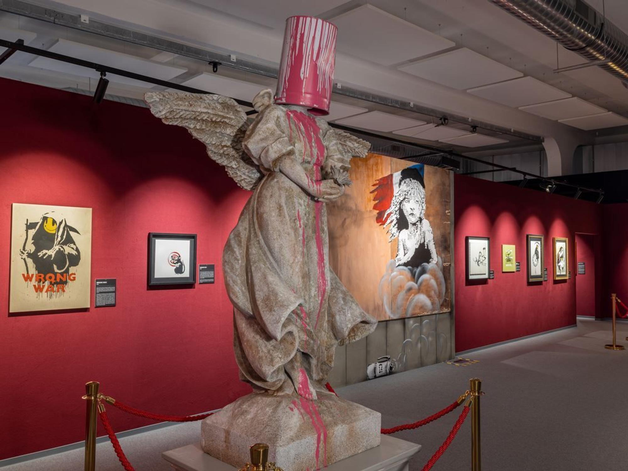 Eine Steinfigur steht mit Eimer auf dem Kopf in einer Kunstausstellung.