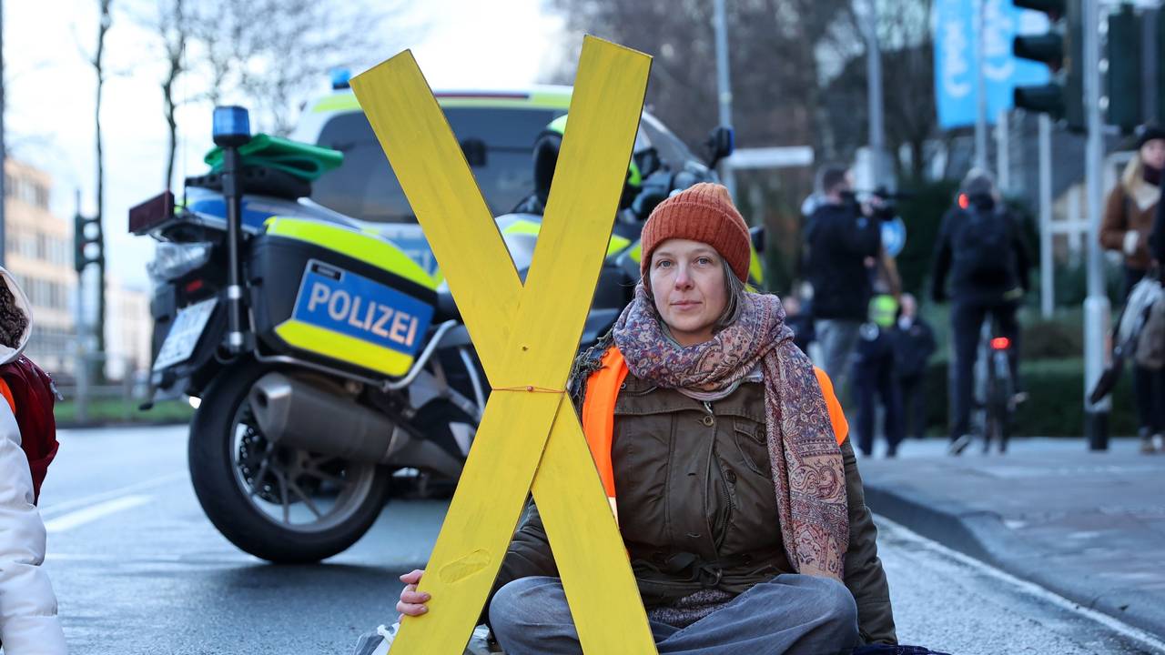 Eine Frau sitzt auf der Fahrbahn und hält ein großes X aus Pappe fest.