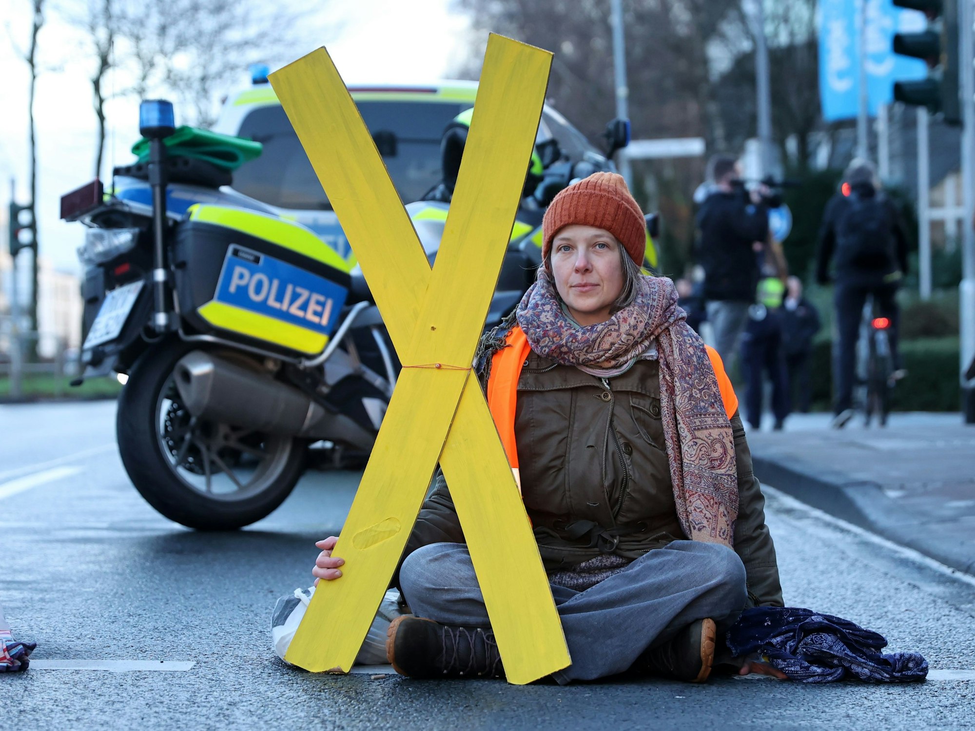Eine Frau sitzt auf der Fahrbahn und hält ein großes X aus Pappe fest.