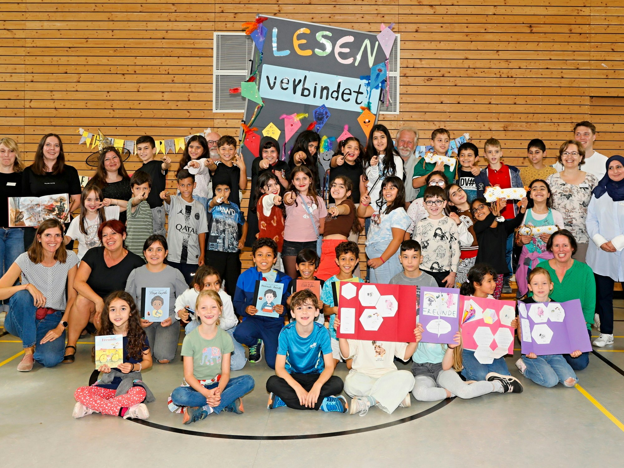 Mehrere Kinder und einige Erwachsene sitzen in einer Sporthalle auf dem Boden.
