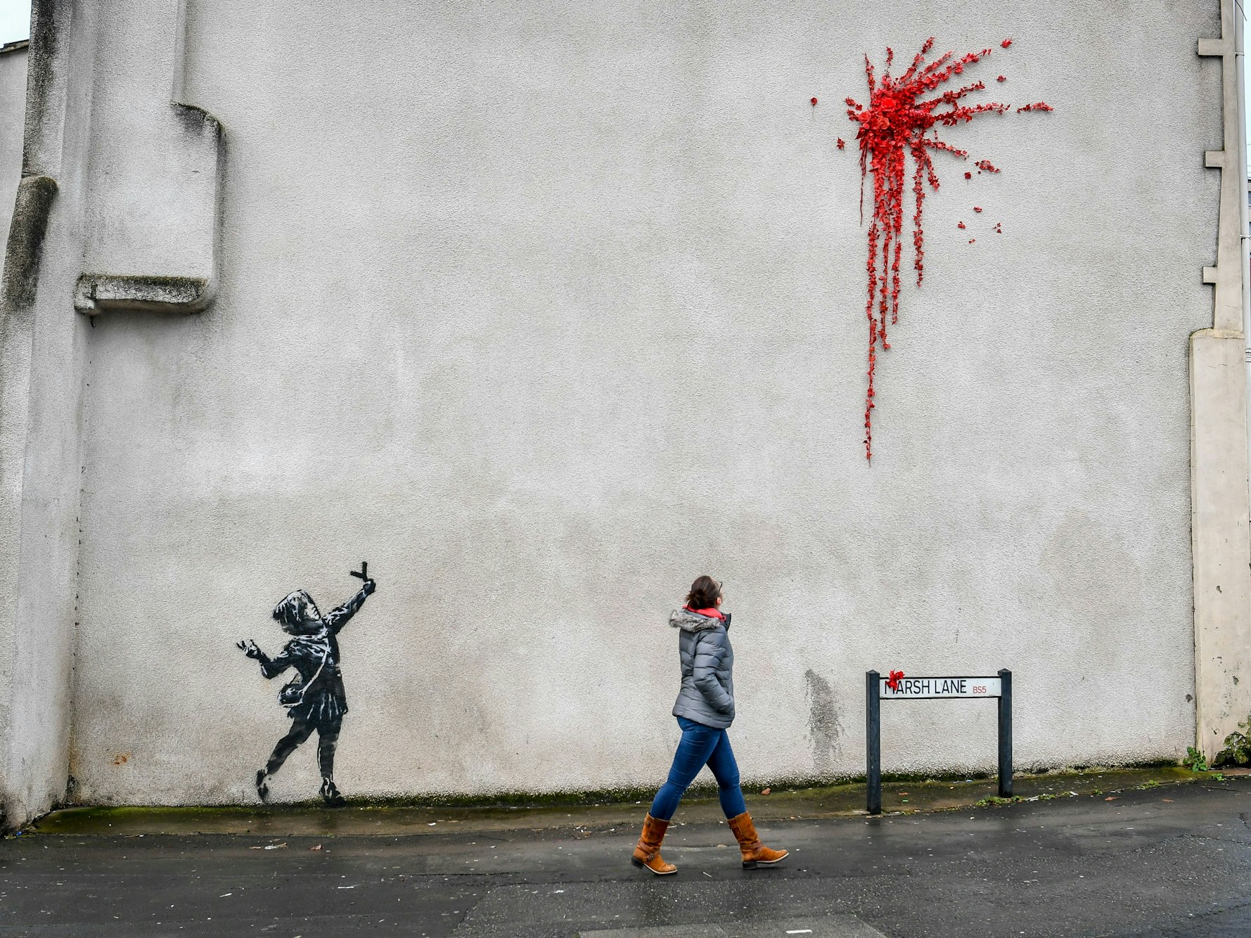 Eine Frau geht an einer Hauswand an einem Street-Art-Werk vorbei, das ein Mädchen mit einer Steinschleuder in der Hand darstellt, über der ein riesiger roter Klecks aus roten Blumen zu sehen ist, hier im Februar 2020 in Bristol.