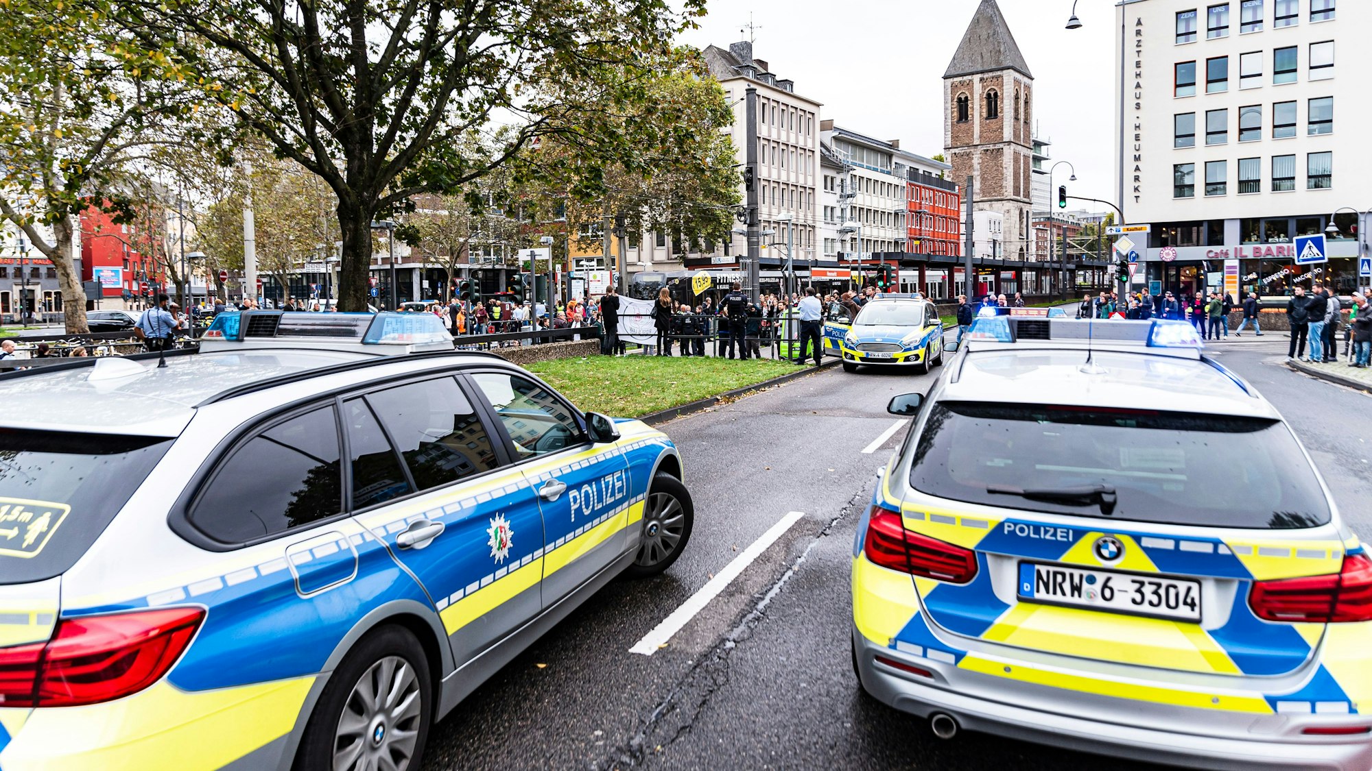 Zwei Polizei-Autos stehen am Kölner Heumarkt, eine Demo im Hintergrund wird überwacht.