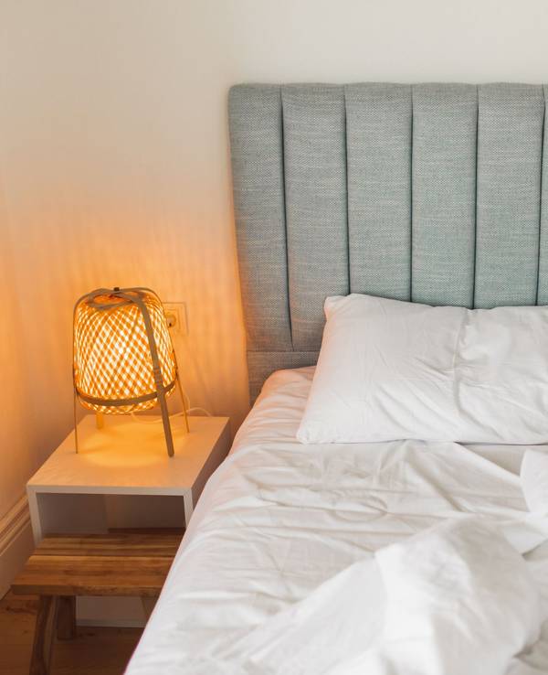 Auf dem Foto sieht man ein graues Bett, links daneben steht ein Nachtschrank mit einer eingeschalteten Lampe darauf.