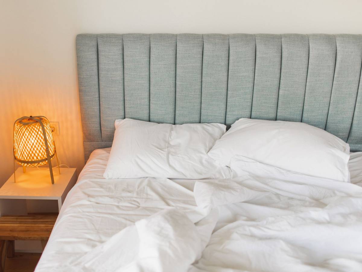 Auf dem Foto sieht man ein graues Bett, links daneben steht ein Nachtschrank mit einer eingeschalteten Lampe darauf.