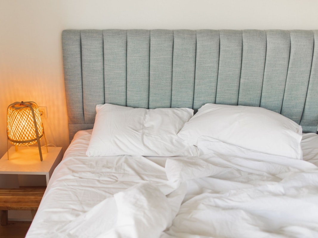 Auf dem Foto sieht man ein graues Bett, links daneben steht ein Nachtschrank mit einer eingeschalteten Lampe darauf.