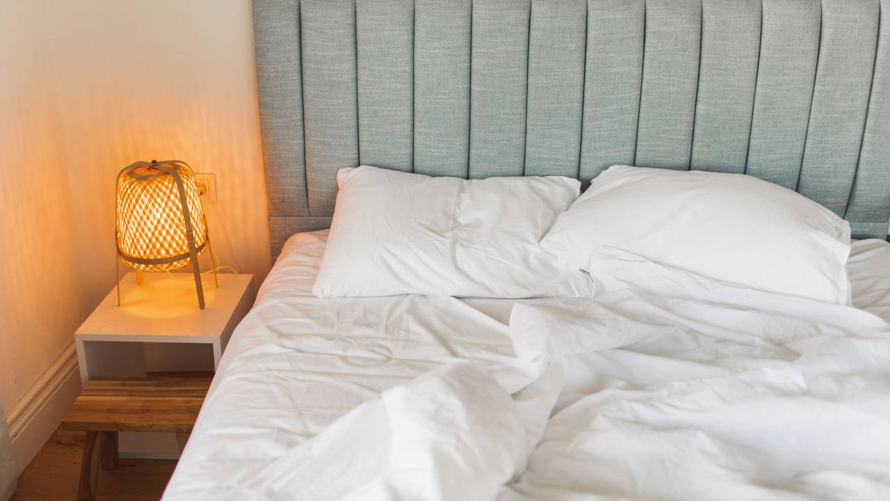 Auf dem Foto sieht man ein graues Bett, links daneben steht ein Nachtschrank mit einer eingeschalteten Lampe darauf.