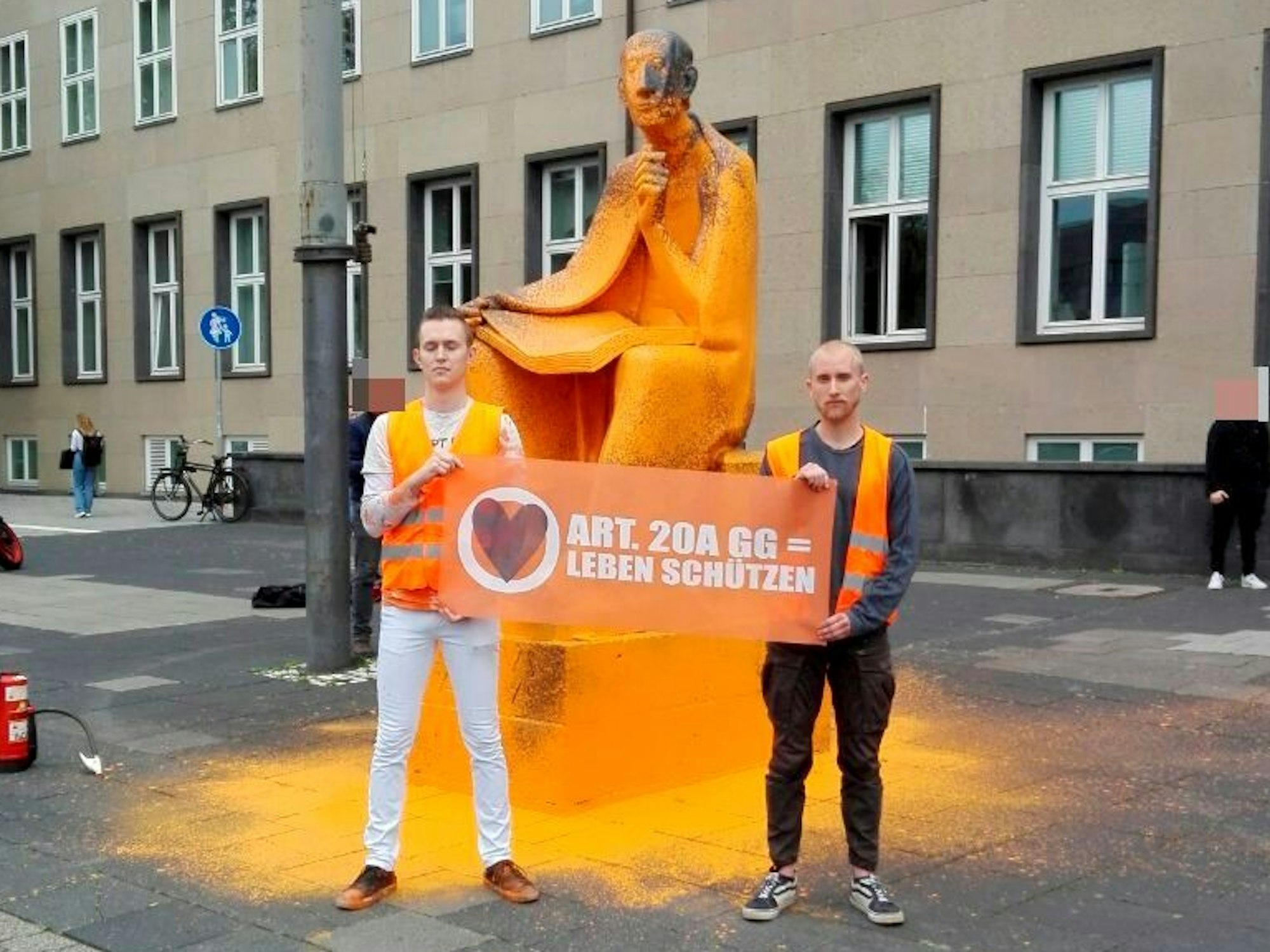 Zwei junge Männer stehen mit einem Plakat vor einem Denkmal, über das Farbe gegossen wurde.
