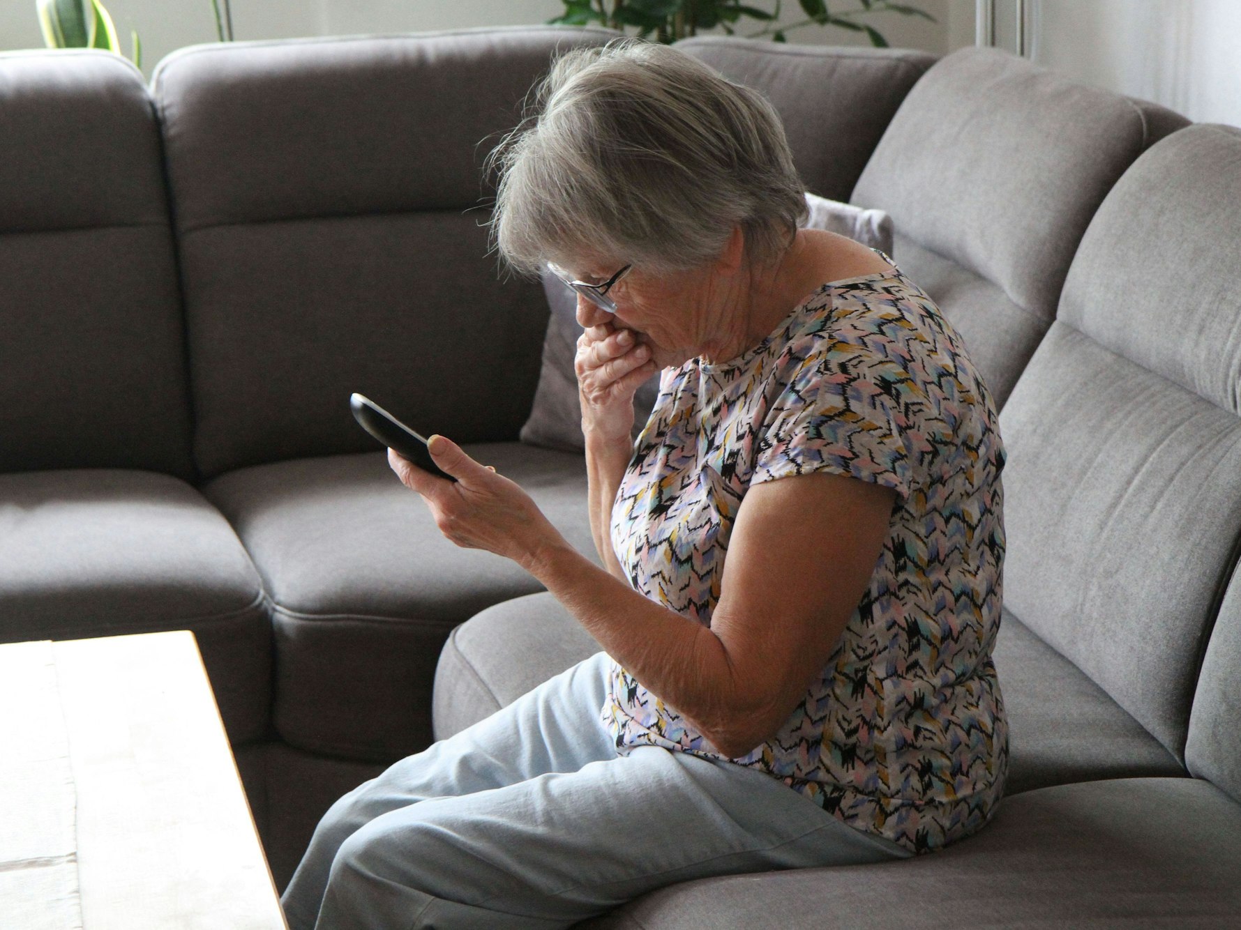 Eine ältere Frau sitzt auf ihrem Sofa und schaut bestürzt auf ihr Handy.