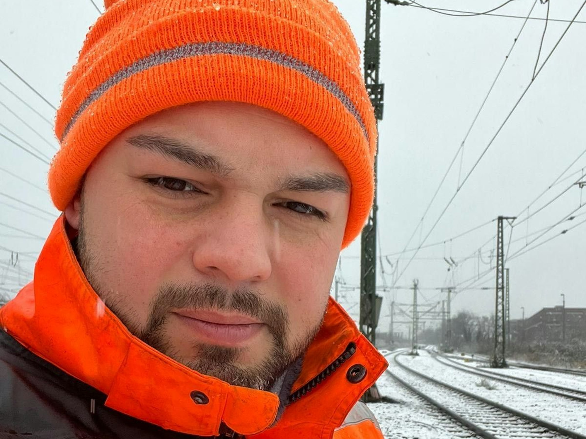 Ein Mann mit Mütze und dicker Jacke steht im Winter nahe Bahngleisen.