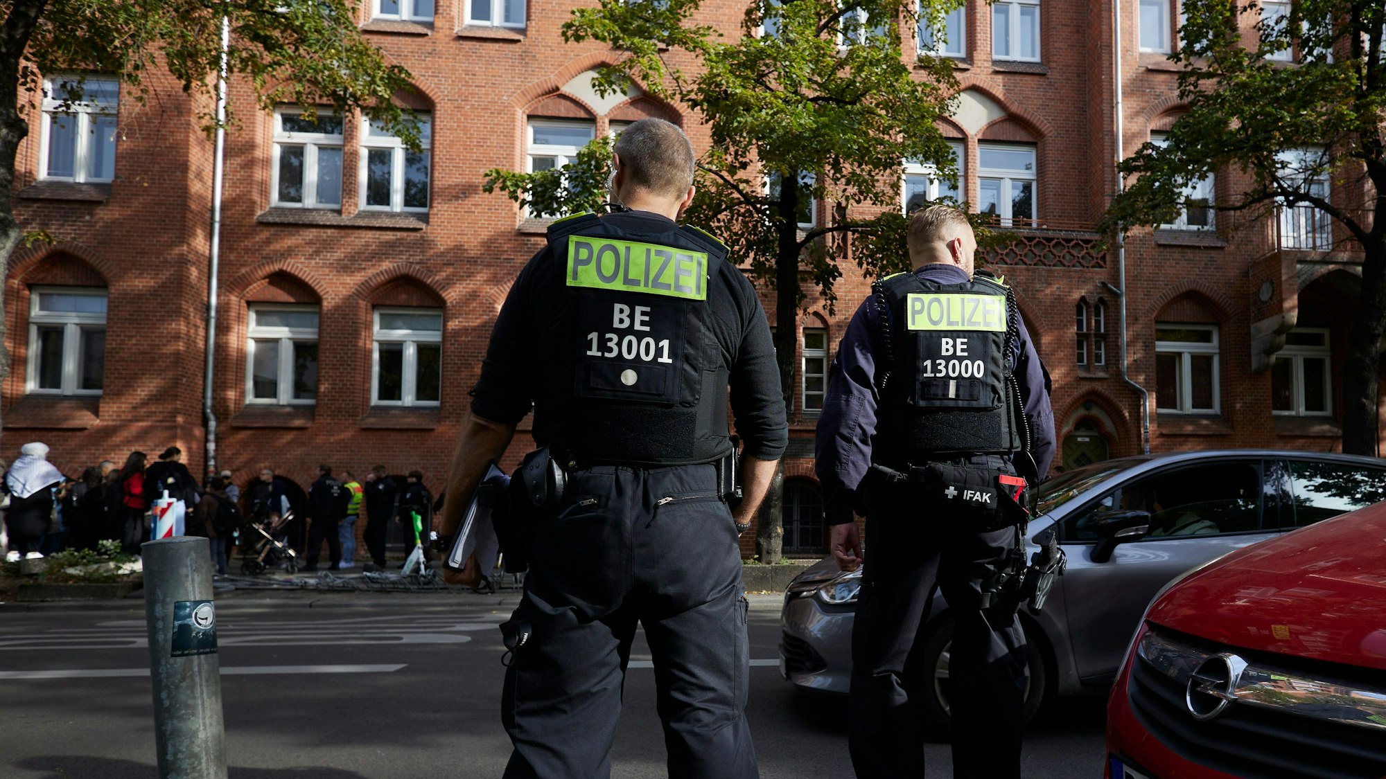 Zwei Polizisten stehen vor einer Berliner Schule. Im Hintergrund sind einige Jugendliche zu sehen.