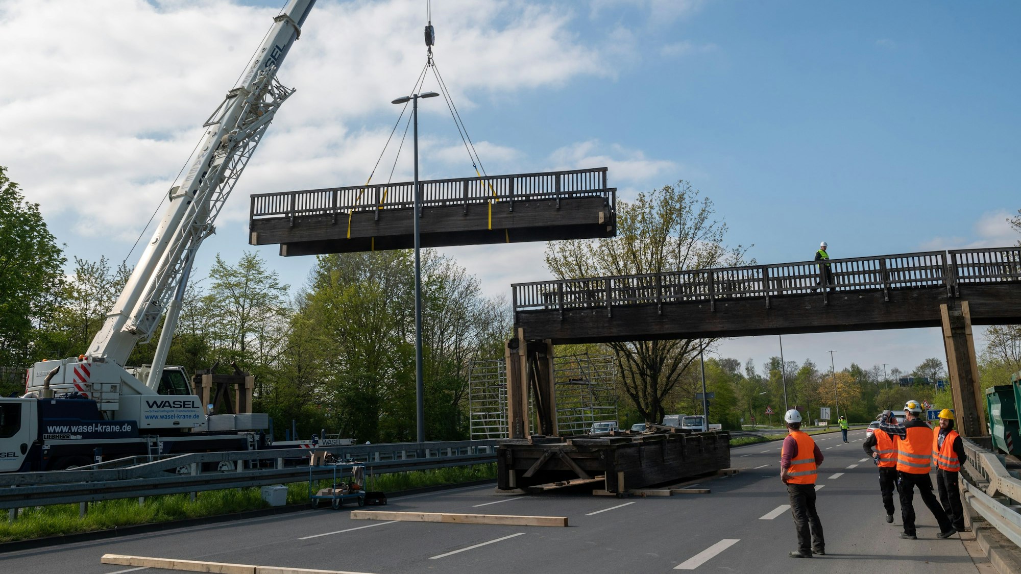 An einem Kran hängt ein Brückenteil. Der Rest der Brücke der über eine mehrspurige Straße führt, steht noch. Auf der Straße befinden sich mehrere Personen in Warnwesten, einige gucken zu dem schwebenden Brückenteil.