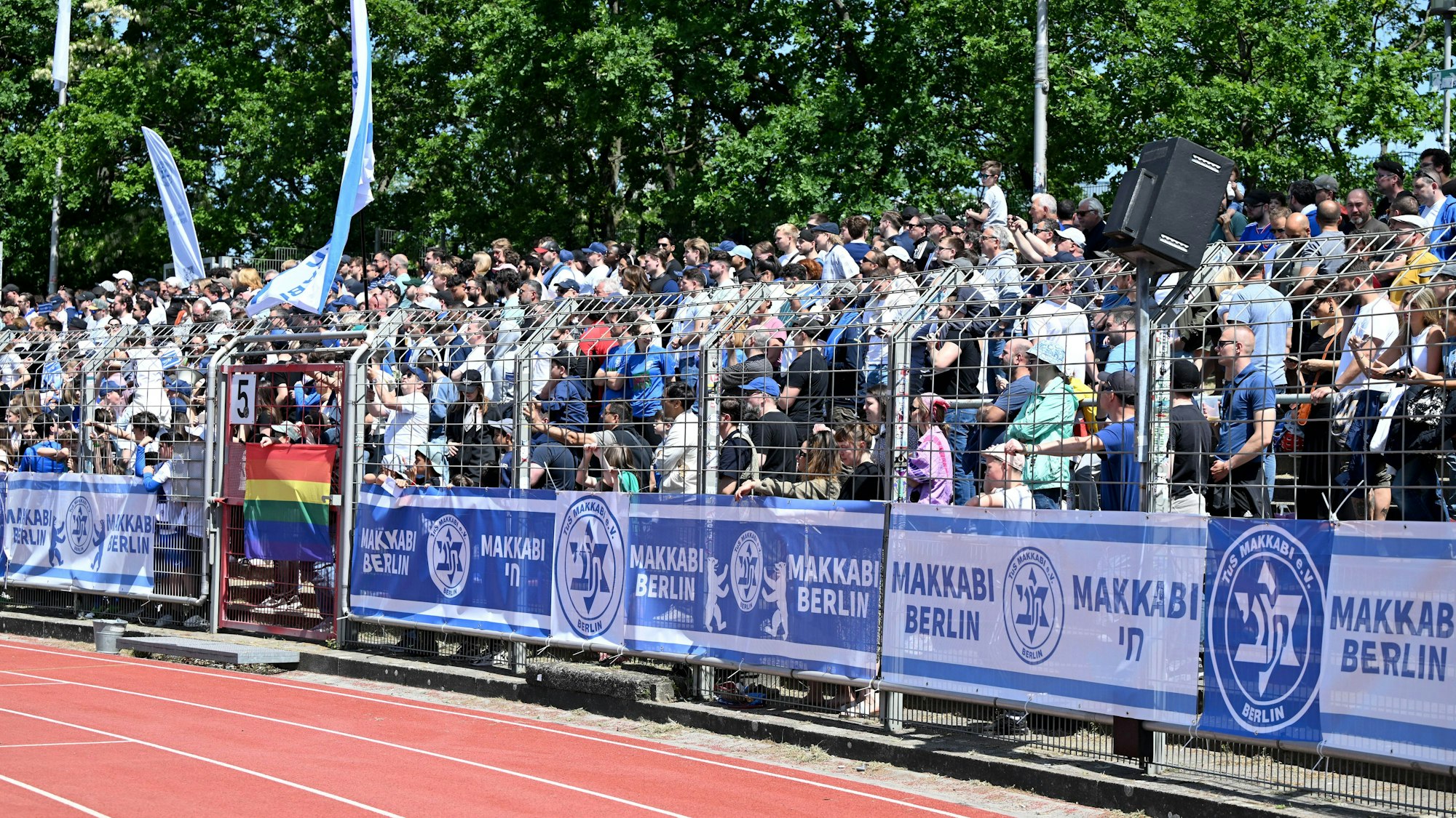 Fußball-Fans eine jüdischen Vereins stehen hinter Zäunen auf einer Stadiontribüne und feuern ihre Mannschaft an.