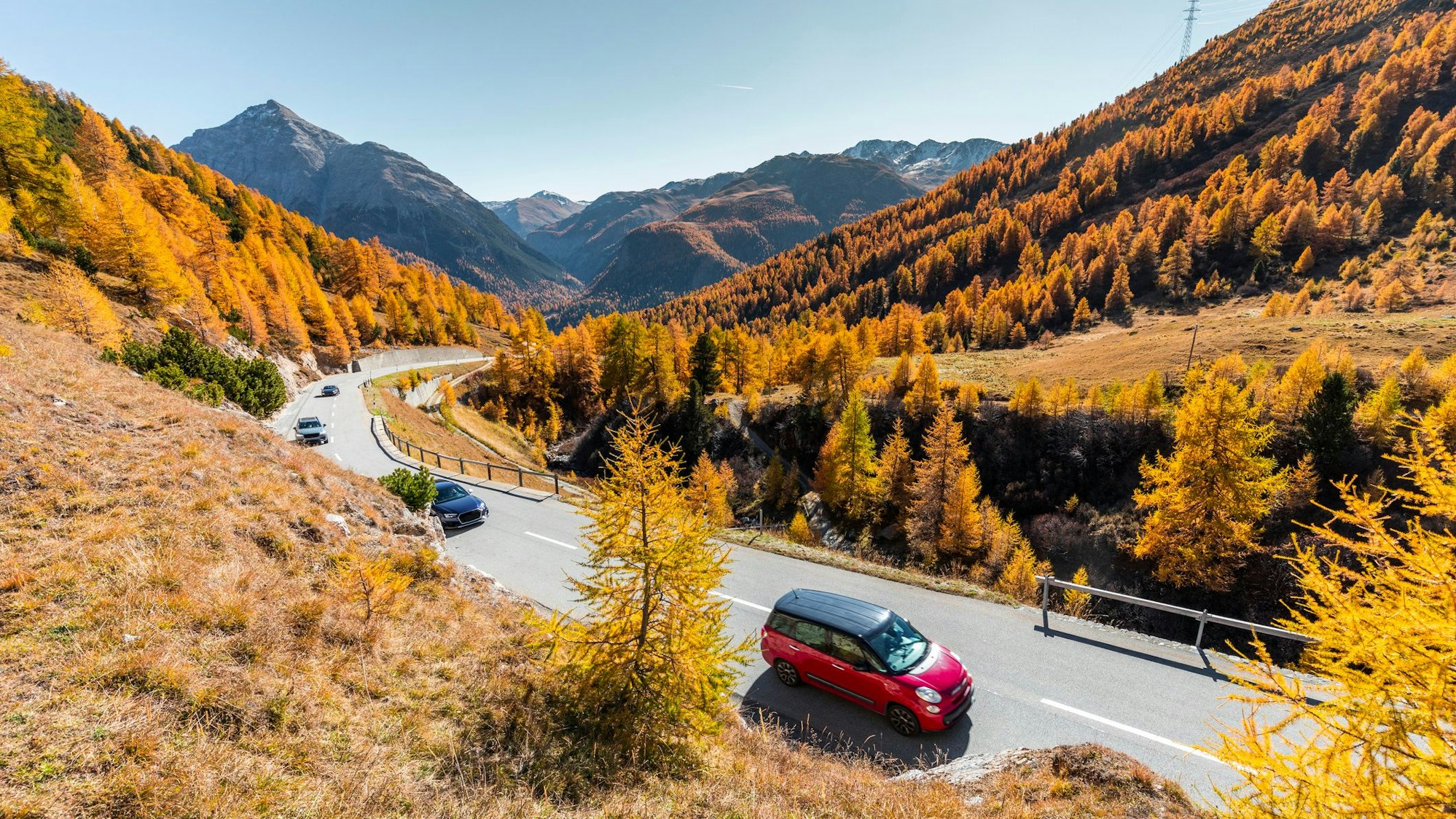 Autos fahren eine Straße zwischen Bergen entlang. Die Blätter der Bäume sind bunt gefärbt.