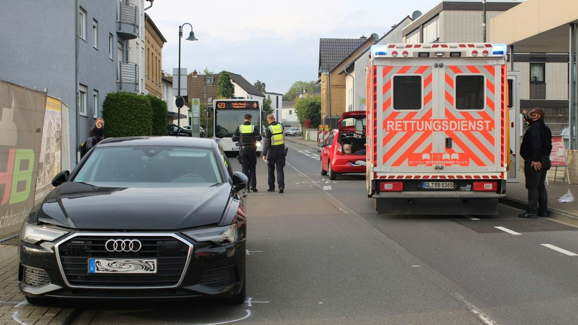 Ein Rettungswagen steht auf der Straße, ebenso zwei Autos, die an einem Unfall beteiligt waren. Zwei Polizisten befinden sich ebenfalls auf der Fahrbahn.