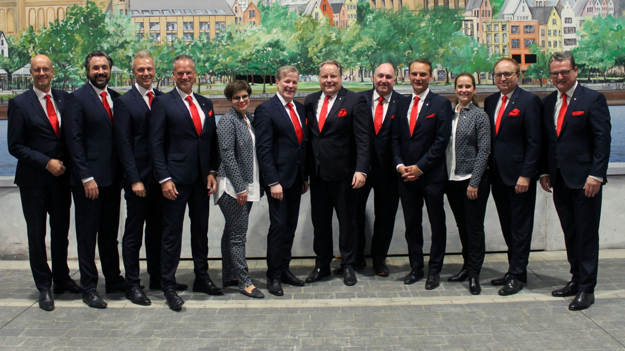Zehn Männer in schwarzen Anzügen und zwei Frauen in grauen Zweiteilern stehen gemeinsam in einer Reihe und schauen in die Kamera.