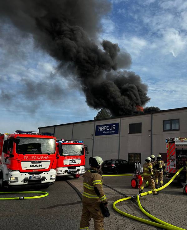 Rauch steigt über einer Lagerhalle auf, davor stehen mehrere Fahrzeuge der Feuerwehr.