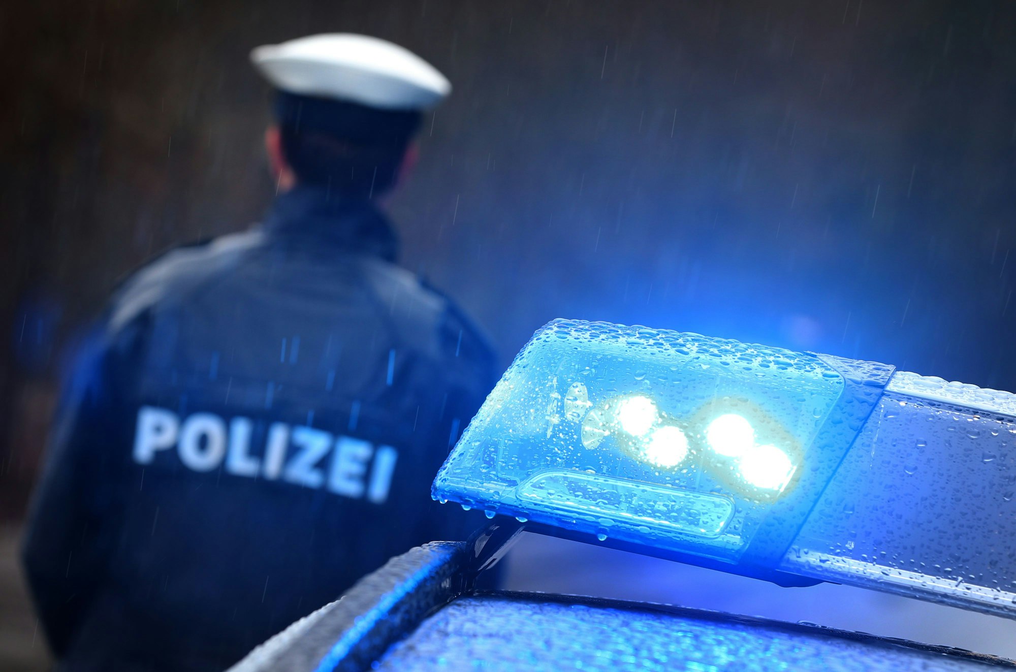 Ein Polizist steht im Regen vor einem Streifenwagen, dessen Blaulicht aktiviert ist.