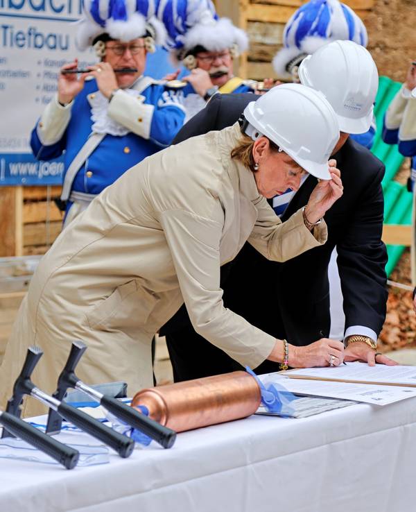 Kölns OB Henriette Reker unterzeichnet auf einer Baustelle eine Urkunde, sie trägt einen weißen Helm.