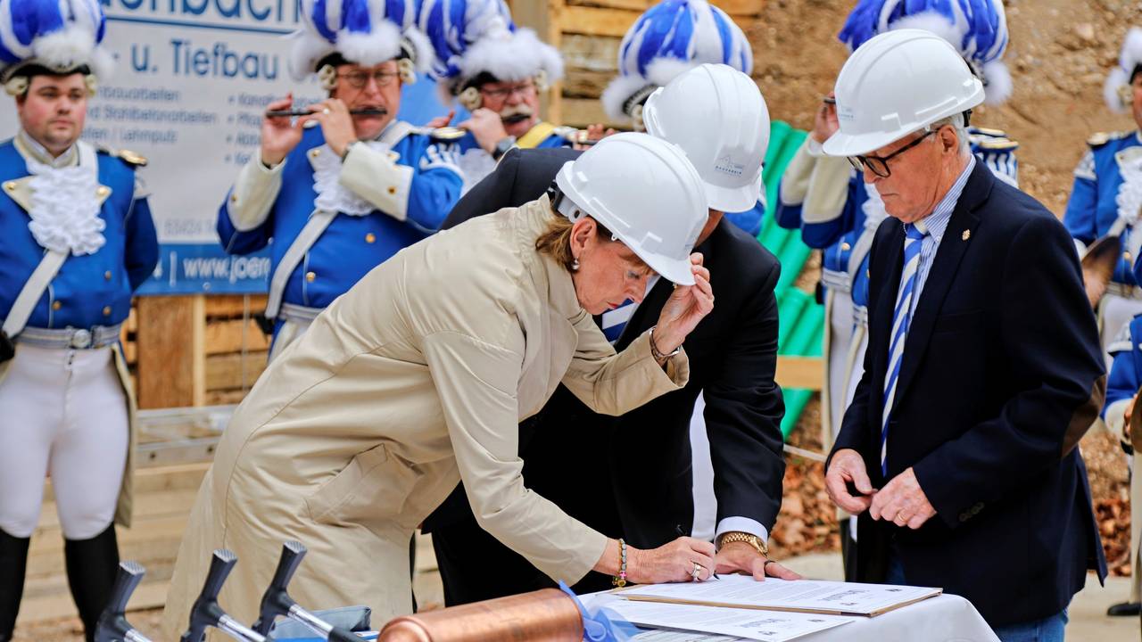 Kölns OB Henriette Reker unterzeichnet auf einer Baustelle eine Urkunde, sie trägt einen weißen Helm.