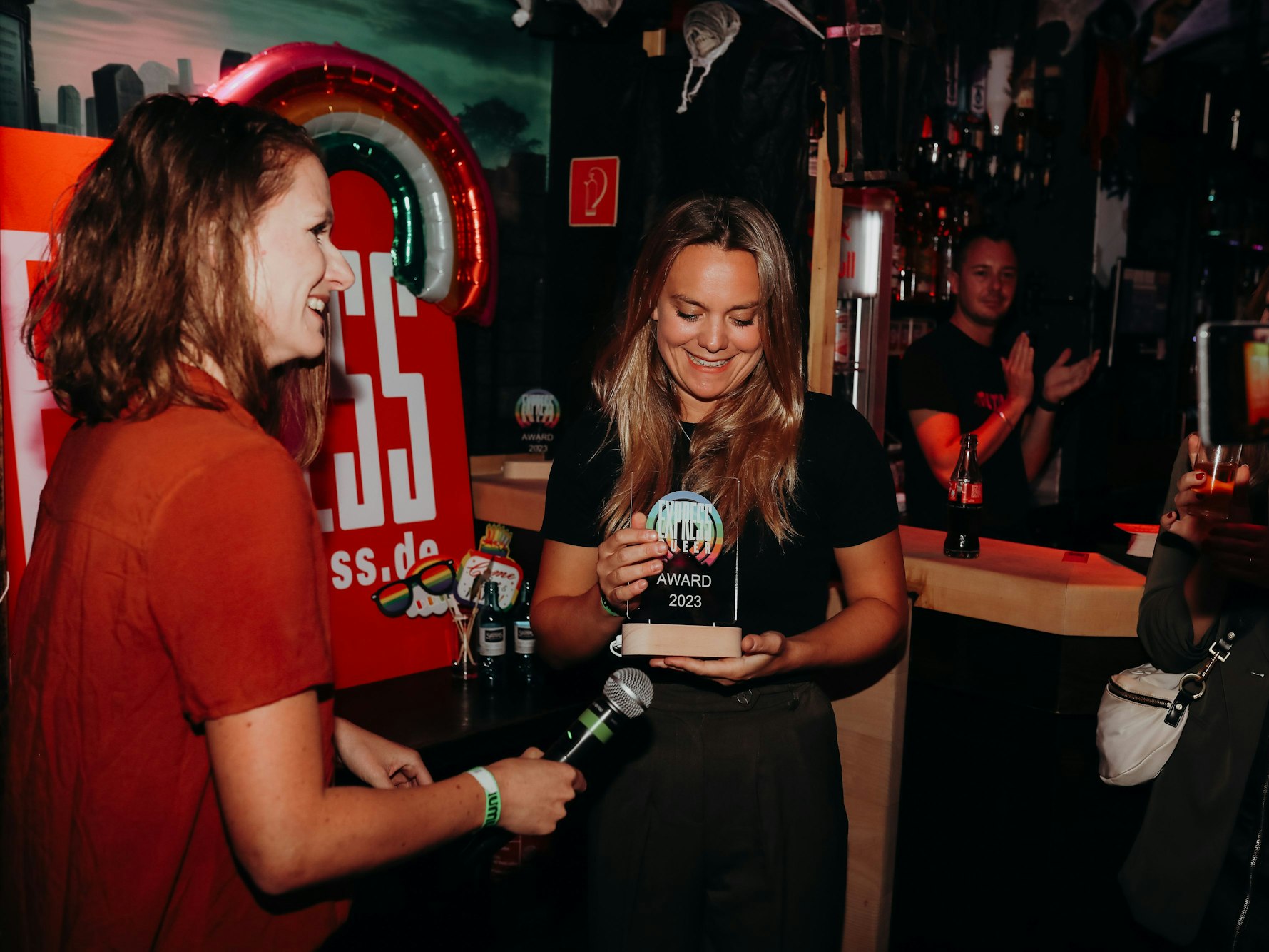 Zwei Frauen gemeinsam auf der Bühne, die eine übergibt der anderen eine Trophäe.