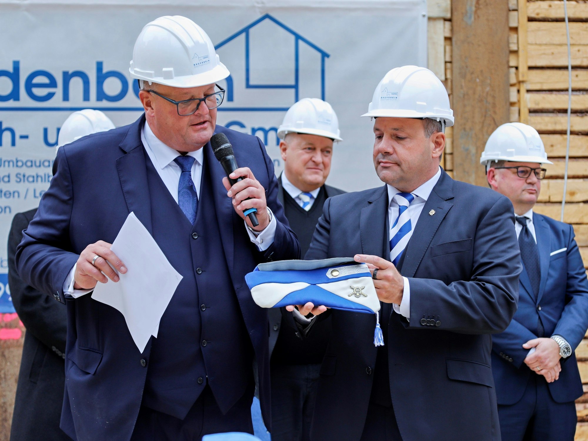 Zwei Männer mit Bauhelmen und in blauen Anzügen legen eine Karnevalskappe ab.
