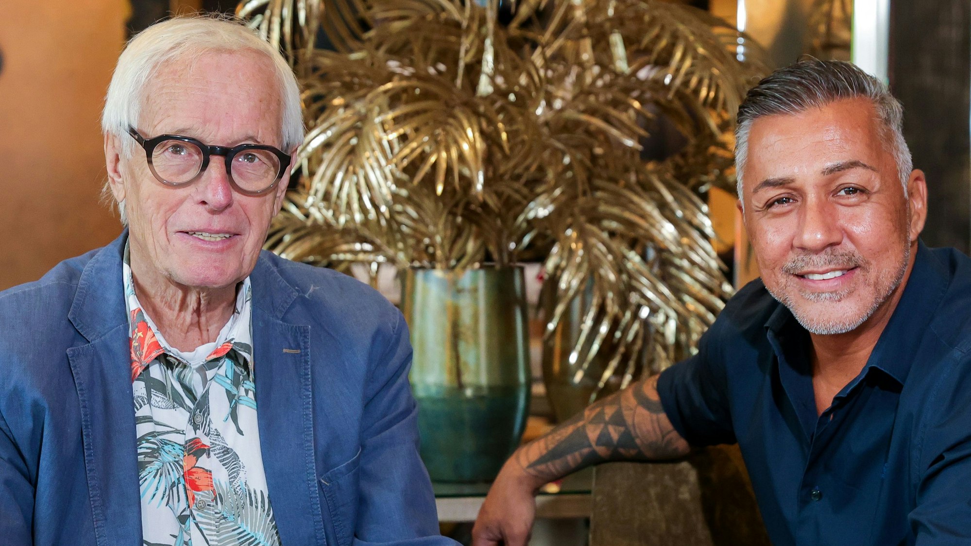 Mike Leon Grosch und EXPRESS-Reporter Horst Stellmacher lächeln beim Gespräch am 31. August 2023 in Köln in die Kamera. Im Hintergrund steht eine goldene Palme.